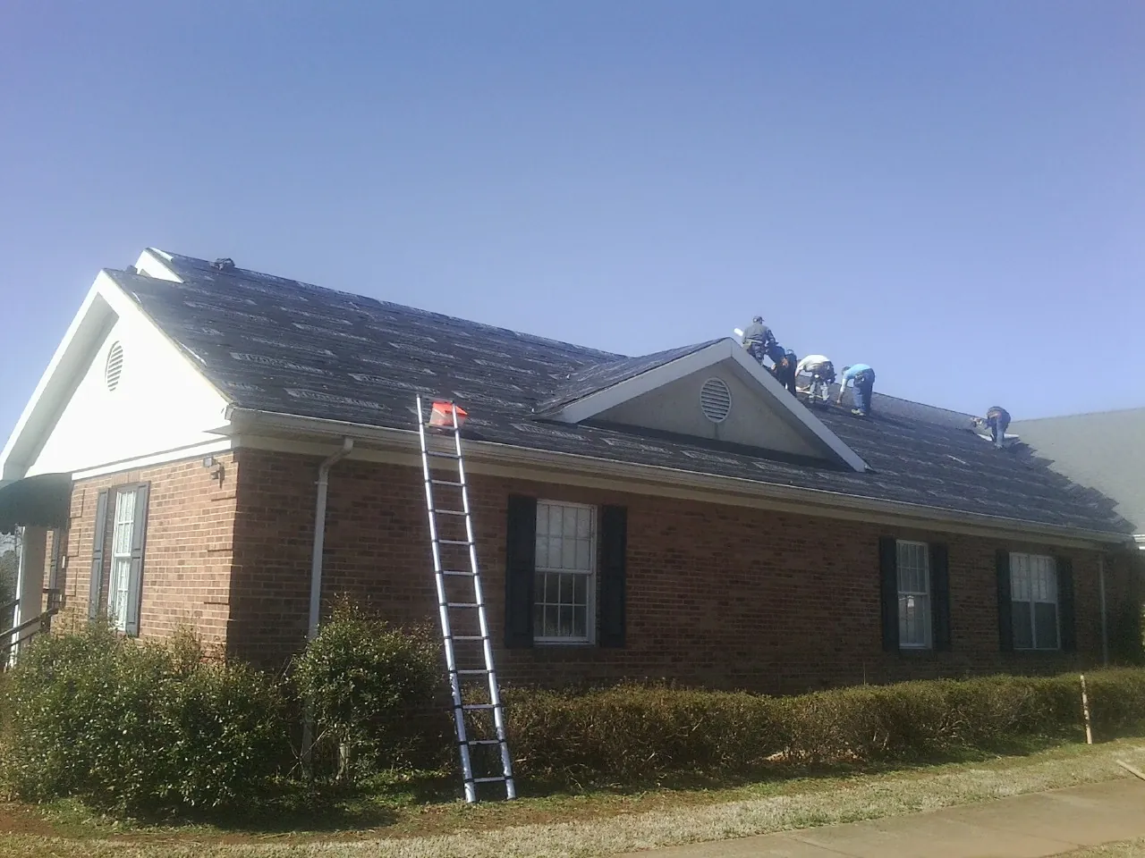 Crew tearing shingles off a brick ranch roof with a ladder set — Emergency Roof Repair work by our crew in Hankinson