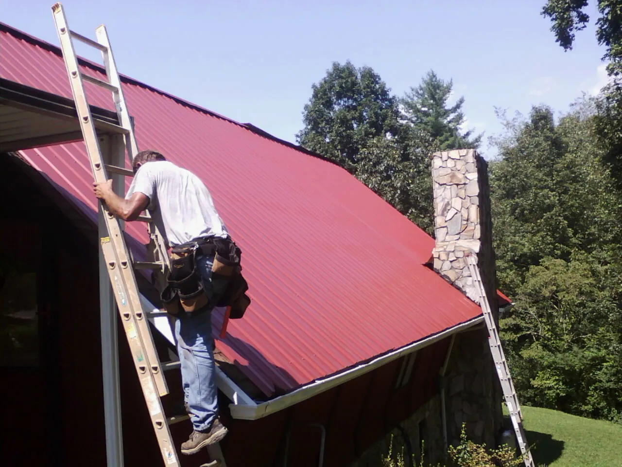 Roofer on a ladder beside a red metal roof and stone chimney — Soffit & Fascia Repair by Roofing Brothers in Hankinson
